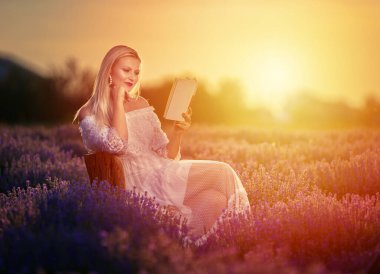 Beyaz elbiseli genç sarışın kadın lavanta tarlasında kitap okuyor.