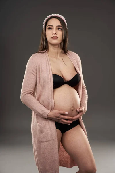 妊娠中の女性で黒ランジェリーとベージュローブポーズ上の灰色の背景 — ストック写真
