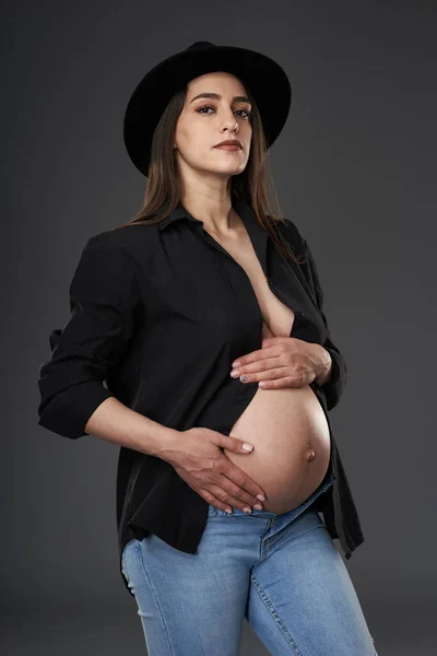 妊娠中の女性で青ジーンズ 帽子とボタンなし黒シャツポージング上の灰色の背景 — ストック写真