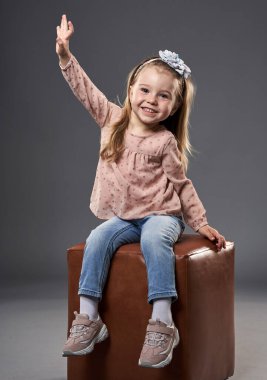 Küçük sevimli kız, gri arka planda izole bir şekilde küp bir sandalyede otururken el sallıyor.