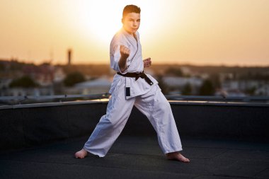 Genç bir çocuk, güneş batarken çatıda beyaz kimono ile antrenman yapıyor.