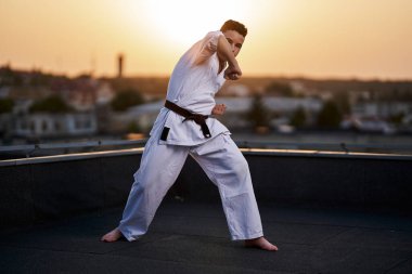 Genç bir çocuk, güneş batarken çatıda beyaz kimono ile antrenman yapıyor.