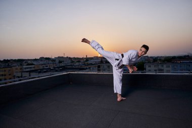 Genç bir çocuk, güneş batarken çatıda beyaz kimono ile antrenman yapıyor.