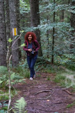 Kıvırcık kızıl saçlı kadın Apuseni dağlarındaki işaretli bir orman patikasında yürüyüş yapıyor. Elinde bir kamera var ve doğal ışık altında yemyeşil ile çevrili.