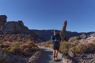 Erkek yürüyüşçü, Teide Ulusal Parkı 'ndaki Roques de Garca' da volkanik kaya oluşumları arasında manzaralı bir patikada yürürken bir aksiyon kamerasıyla vlog yapıyor. 