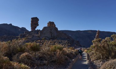 Erkek yürüyüşçü, Teide Ulusal Parkı 'ndaki Roques de Garca' da volkanik kaya oluşumları arasında manzaralı bir patikada yürürken bir aksiyon kamerasıyla vlog yapıyor. 