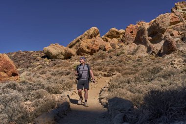 Yanardağ oluşumları ve Teide Ulusal Parkı 'nda açık mavi gökyüzünün altında kuru çalılarla çevrili kayalık patika boyunca yürüyen kamera ekipmanları taşıyan erkek yürüyüşçü.