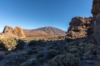 Tenerife, Teide Ulusal Parkı 'ndaki Roques de Garcia' nın engebeli volkanik oluşumları ve kuru bitkileri tarafından çerçevelenmiş gün batımında görülen Teide Dağı.