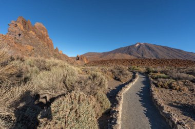 Teide Dağı 'na doğru giden kavisli yürüyüş yolu ve Tenerife' deki Teide Ulusal Parkı 'nda mavi gökyüzünün altında Roques de Garcia' nın volkanik kaya oluşumları.
