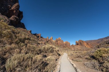 Volkanik kaya oluşumu, Teide Ulusal Parkı, Tenerife 'deki Roques de Garcia' da açık mavi gökyüzü altında bir yürüyüş yolu ve çöl bitkisi üzerinde yükseliyor.
