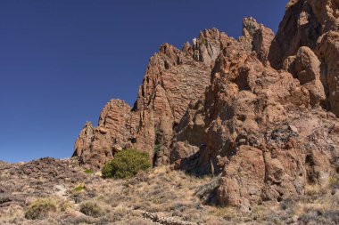 Tenerife 'deki Roques de Garcia' nın kurak ön planında seyrek yeşil çalılar bulunan açık mavi gökyüzüne karşı yükselen yüksek kırmızı kaya oluşumları