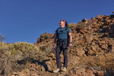 Parlak sabah güneşi altında kayalık bir volkanik patikada duran gülümseyen erkek yürüyüşçü Teide Dağı yakınlarındaki yamaçlara doğru bakıyor.