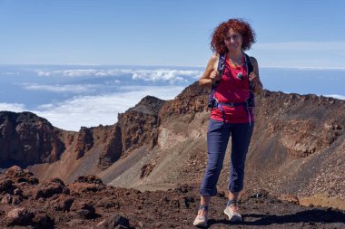 Pico Viejo kraterinin kenarında gülümseyen kadın yürüyüşçü berrak gökyüzünün altında, engebeli volkanik kayalıklar ve lav oluşumları arkasında