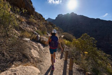 Kasklı ve sırt çantalı kadın yürüyüşçü Tenerife 'deki Barranco del Infierno kanyonunda güneşli patika boyunca yürüyor
