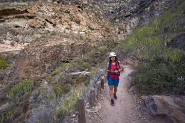 Kasklı ve kameralı kadın doğa fotoğrafçısı Barranco del Infierno volkanik kanyonu boyunca dar patika boyunca yürüyor.