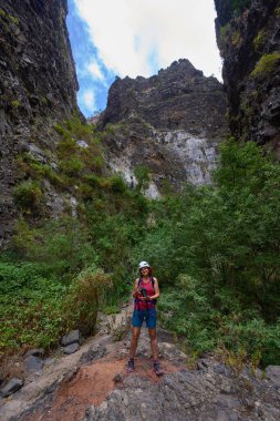 Gülümseyen kadın fotoğrafçı, Barranco del Infierno 'daki volkanik kanyon duvarlarının altındaki yemyeşil bitki örtüsüyle çevrili kayalık yolda gururla duruyor.