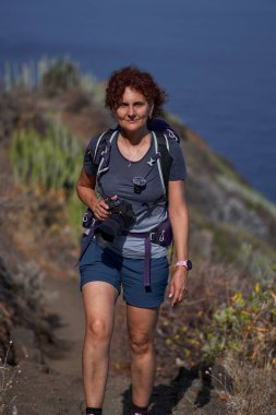 Kameralı ve sırt çantalı bir kadın yürüyüşçü Atlantik Okyanusu 'nun arka planıyla Taganana' ya doğru yürüyor.