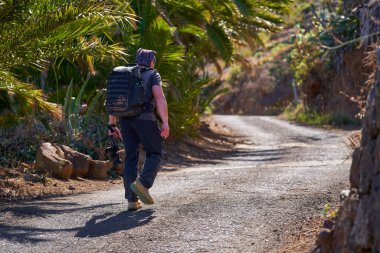 Sırt çantalı ve kameralı bir erkek fotoğrafçı Anaga bölgesindeki palmiye ağaçlarıyla dolu kırsal yolda yokuş yukarı yürüyor.