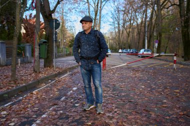 Sırt çantalı orta yaşlı bir adam sonbahar caddesinde park girişinin yanındaki yapraklarla kaplı yürüyor.