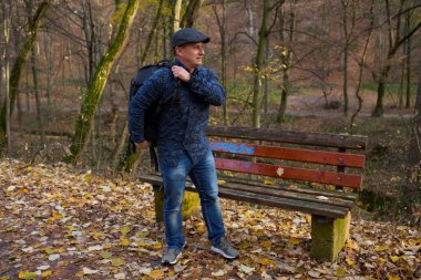 Orta yaşlı bir adam orman parkında yapraklarla kaplı ahşap bankın yanında sırt çantasını kaldırıyor.