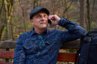 Orta yaşlı bir adam ahşap parktaki bankta oturuyor ve sonbahar ormanında yanında sırt çantasıyla akıllı telefonuyla konuşuyor.