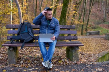 Orta yaşlı dijital göçebe sonbahar ormanlarında park bankında otururken tablet üzerinde çalışıyor.