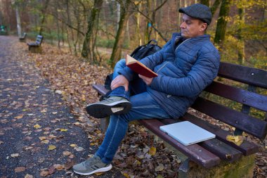 Orta yaşlı bir adam, sonbahar ormanında yanında dizüstü bilgisayarla parkta oturmuş kitap okuyor.