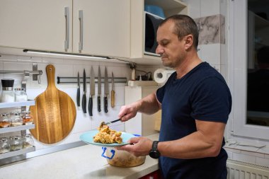 Lacivert tişörtlü, odaklanmış olgun bir adam modern, parlak bir mutfakta tabaklara yemek servisi yapıyor.