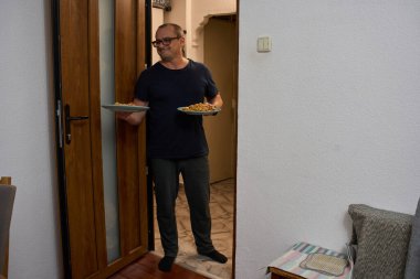 Donanma mavisi tişörtlü ve gözlüklü gülümseyen bir adam kapıda duruyor ve odaya iki tabak lahana güveci taşıyor.