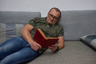 Tişört ve kot giyen bir adam kanepeye oturmuş, ayakları havada, elinde kırmızı bir kitap tutuyor ve okuyor.