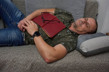 Tişört ve kot giyen bir adam kanepeye uzanmış, gözleri kapalı, kırmızı bir kitap okurken uyuyakalmış.