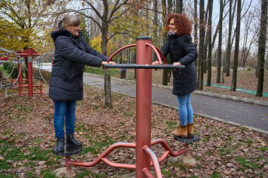 İki bayan arkadaş, sonbahar parkında birlikte spor aletleri kullanarak eğleniyorlar.