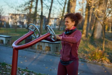 Atletik kadın, sonbahar parkında yaprakları dökülmüş bir spor salonunda eliptik çapraz antrenör üzerinde egzersiz yapıyor.