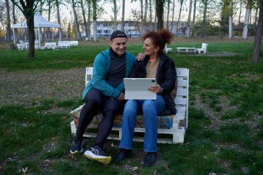 Kadın tablette gülümseyen adama fotoğraf gösterirken, bir çift parkta beyaz palet bankında oturuyor.