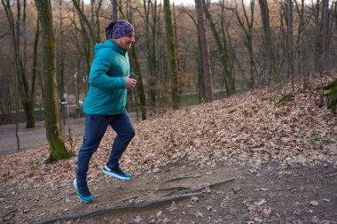 Orta yaşlı, deniz mavisi ceketli bir adam elinde akıllı bir telefonla sonbahar ormanında yaprak kaplı patikada yokuş yukarı koşuyor.