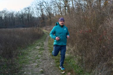 Gök mavisi ceketli orta yaşlı bir adam sonbahar çayırlarında çıplak ağaçlarla dar patika boyunca koşuyor.