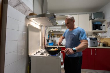 Orta yaşlı, mavi gömlekli bir adam mutfaktaki gaz sobasında tavada sebzelerle makarna karıştırıyor.