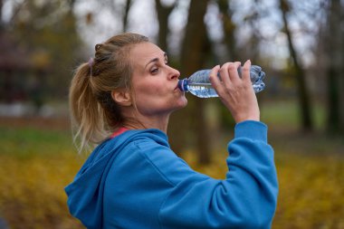 Sonbahar parkında koştuktan sonra şişeden su içen orta yaşlı bir kadın.