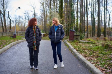 Sonbaharda bir park yolunda yürüyen ve konuşan iki orta yaşlı kadın arkadaş.
