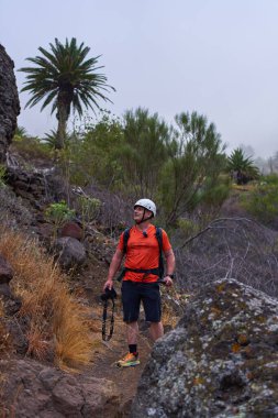 Kayalık Masca geçidinde kaskı ve kamerası olan erkek yürüyüşçü palmiye ağaçları ve volkanik kayalarla çevrili.