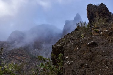 Yoğun atmosferik sis altında seyrek yeşil bitki örtüsü ve dik uçurumlar içeren engebeli volkanik yamaçlar..
