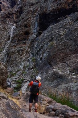 Kasklı ve sırt çantalı bir erkek fotoğrafçı yürüyüş parkurundaki yüksek Masca vadisi uçurumlarının fotoğraflarını çekiyor.