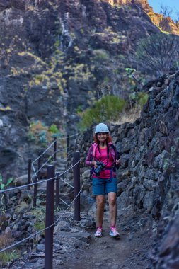 Kamera ve kaskla gülümseyen kadın yürüyüşçü volkanik kaya duvarları ve parmaklıklar arasında Masca geçidinde yürüyor.