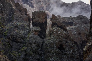 Büyük dikey volkanik kaya duvarı, mağara açıkları ve sisli Masca geçidi ile kaplı yosun yüzeyi.