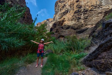 Kollarını açıp Masca vadisinde zıplayan kadın yürüyüşçü. Etrafı yemyeşil bitki örtüsü ve yüksek uçurumlarla çevrili.