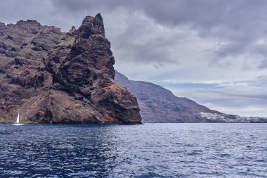 Yelkenli Los Gigantes volkanik kayalıklarından geçiyor. Uzak bir kasaba ve Tenerife 'deki bulutlu gökyüzünün altında liman.