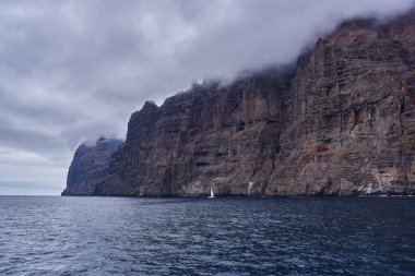 Tenerife 'de bulutlu gökyüzünün altında bitki örtüsü olan Los Gigantes volkanik kayalıklarının yakınına demirlemiş yelkenli ve katamaran gündüz görüşü