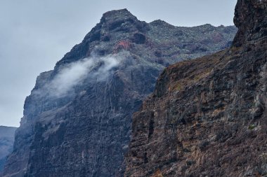 Gri gökyüzünün altındaki katmanlı volkanik kaya oluşumları boyunca sürüklenen sis ile yükselen Masca vadisi uçurumları