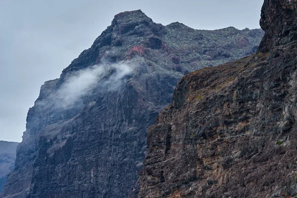 Gri gökyüzünün altındaki katmanlı volkanik kaya oluşumları boyunca sürüklenen sis ile yükselen Masca vadisi uçurumları