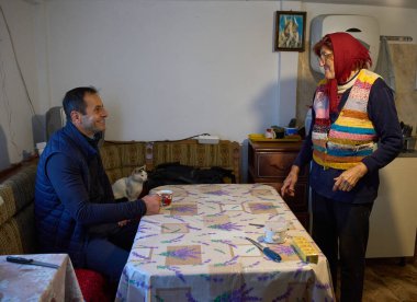 Orta yaşlı bir adam kırsal evdeki mutfak masasında yaşlı bir anneyle çay içiyor.